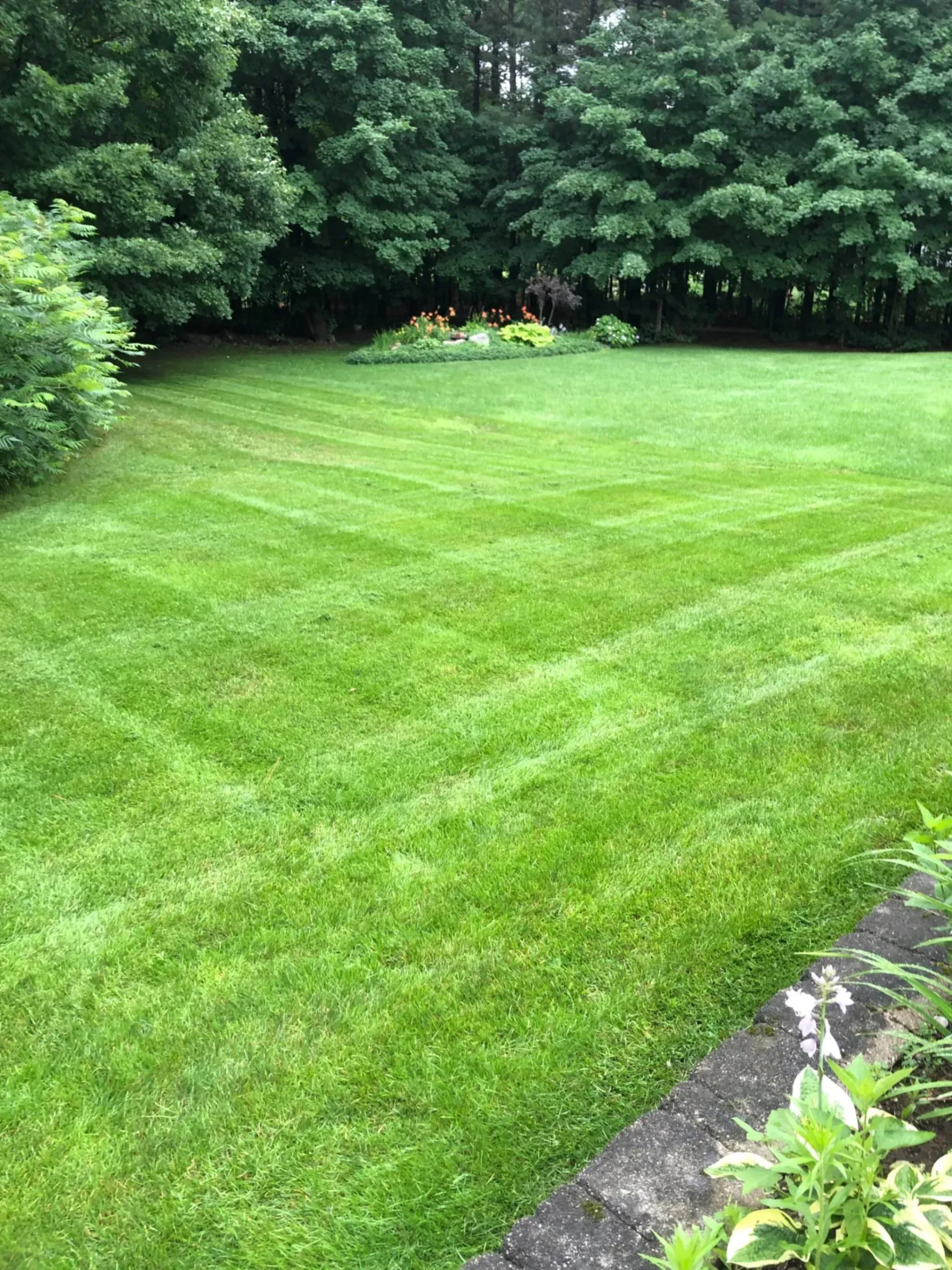 Green lawn with diagonal mowing lines; trees in the background, a small garden visible.