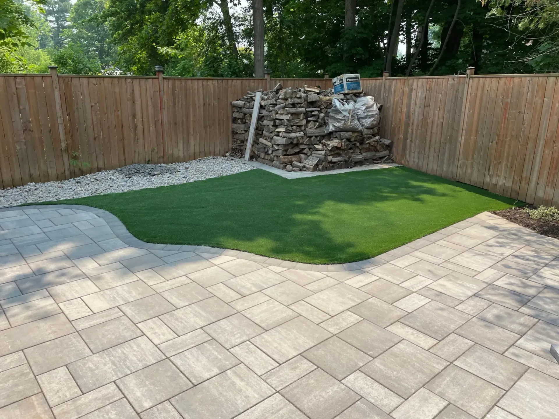 Backyard with a stone patio, artificial grass, wood fence, and stacked firewood.