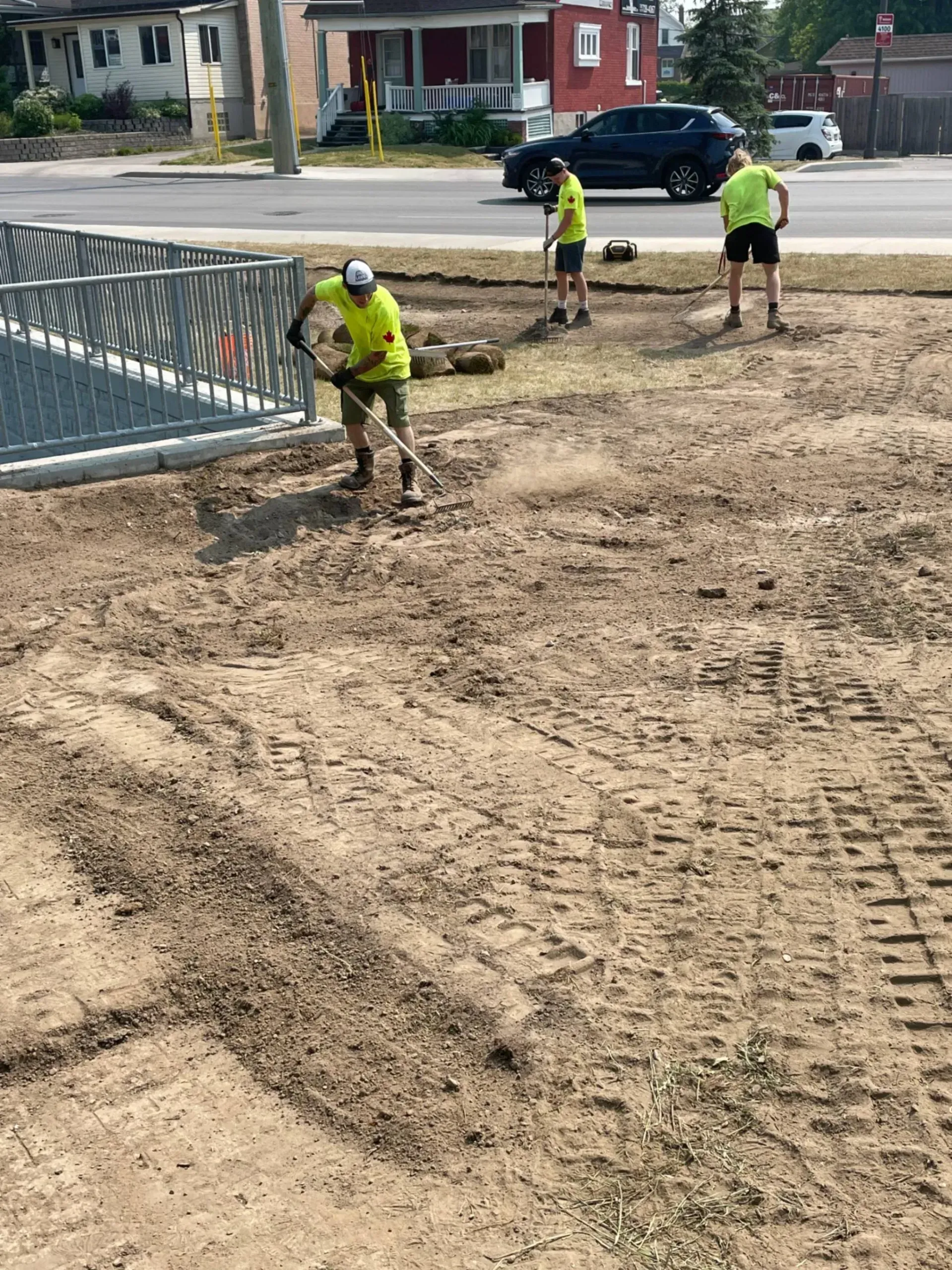 Three workers in neon vests raking dirt, near a fence and road.