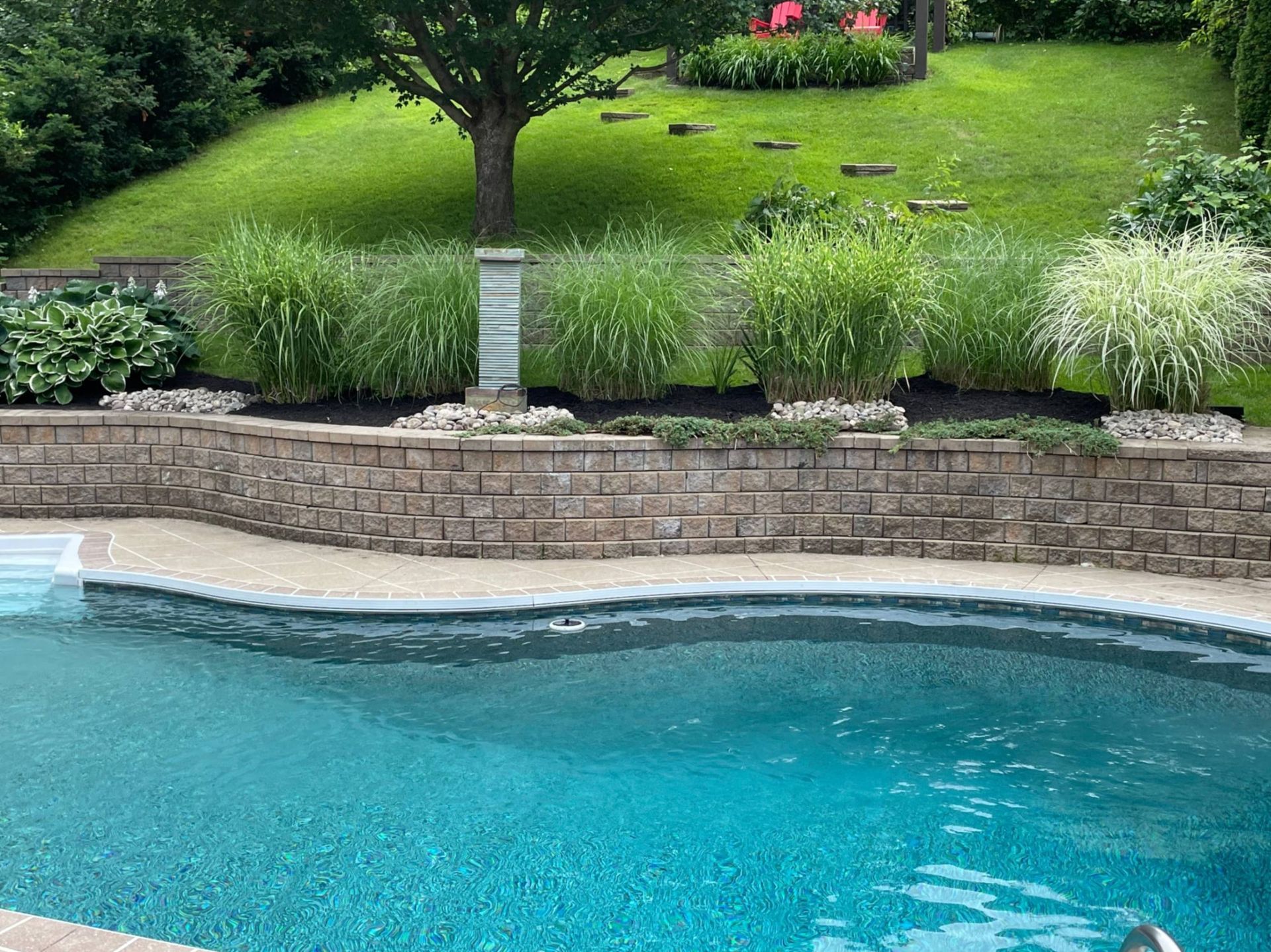 Poolside landscape: brick retaining wall, ornamental grasses, sloping green lawn, and a turquoise pool.