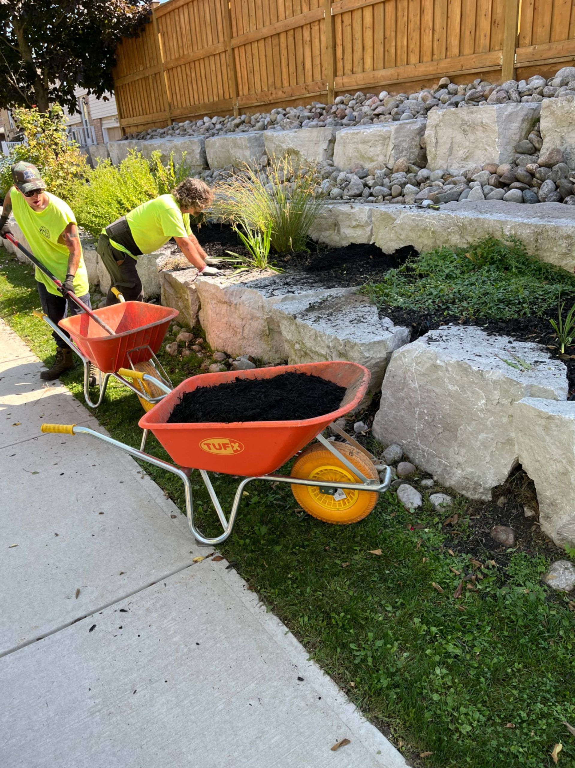 Two people in green shirts spread mulch with wheelbarrows along a rock wall.