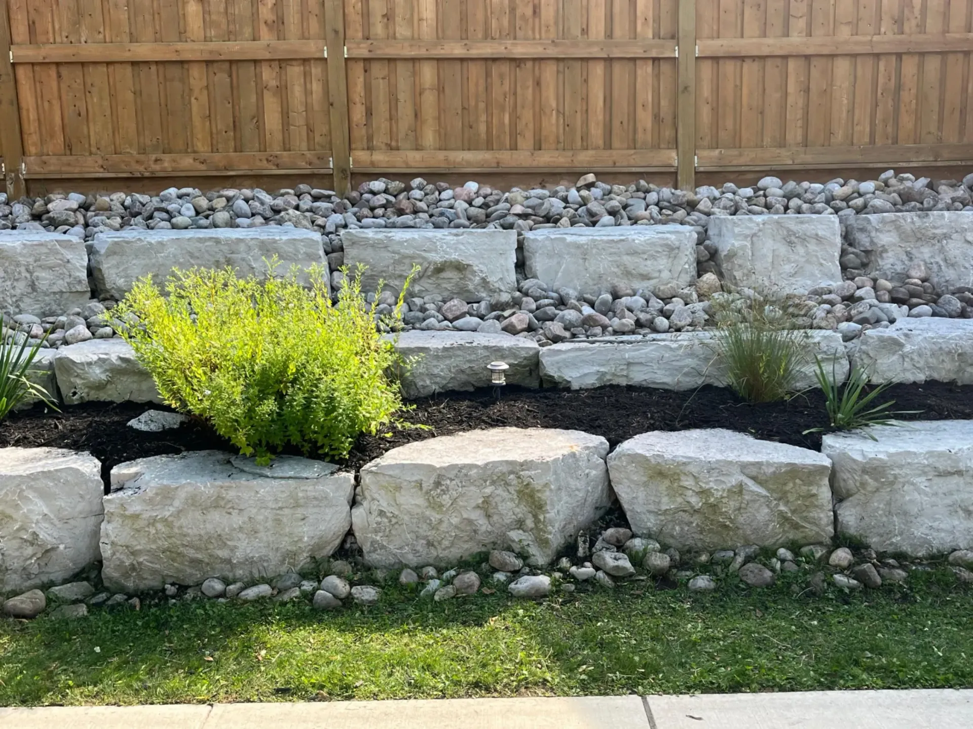 Stone retaining wall with plants, rocks, and a wooden fence backdrop.