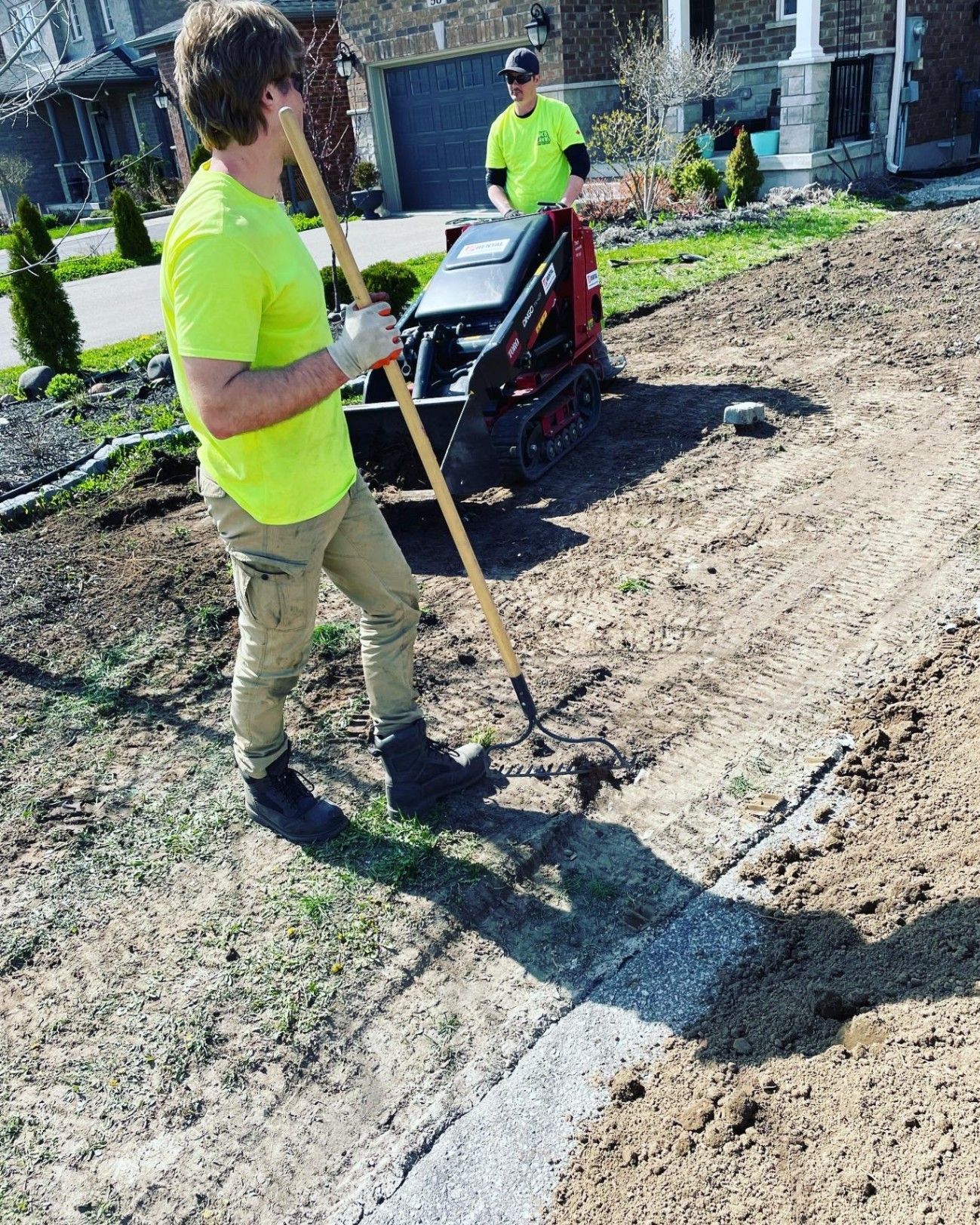 Two workers raking and operating a mini-digger in a yard, prepping for landscaping.