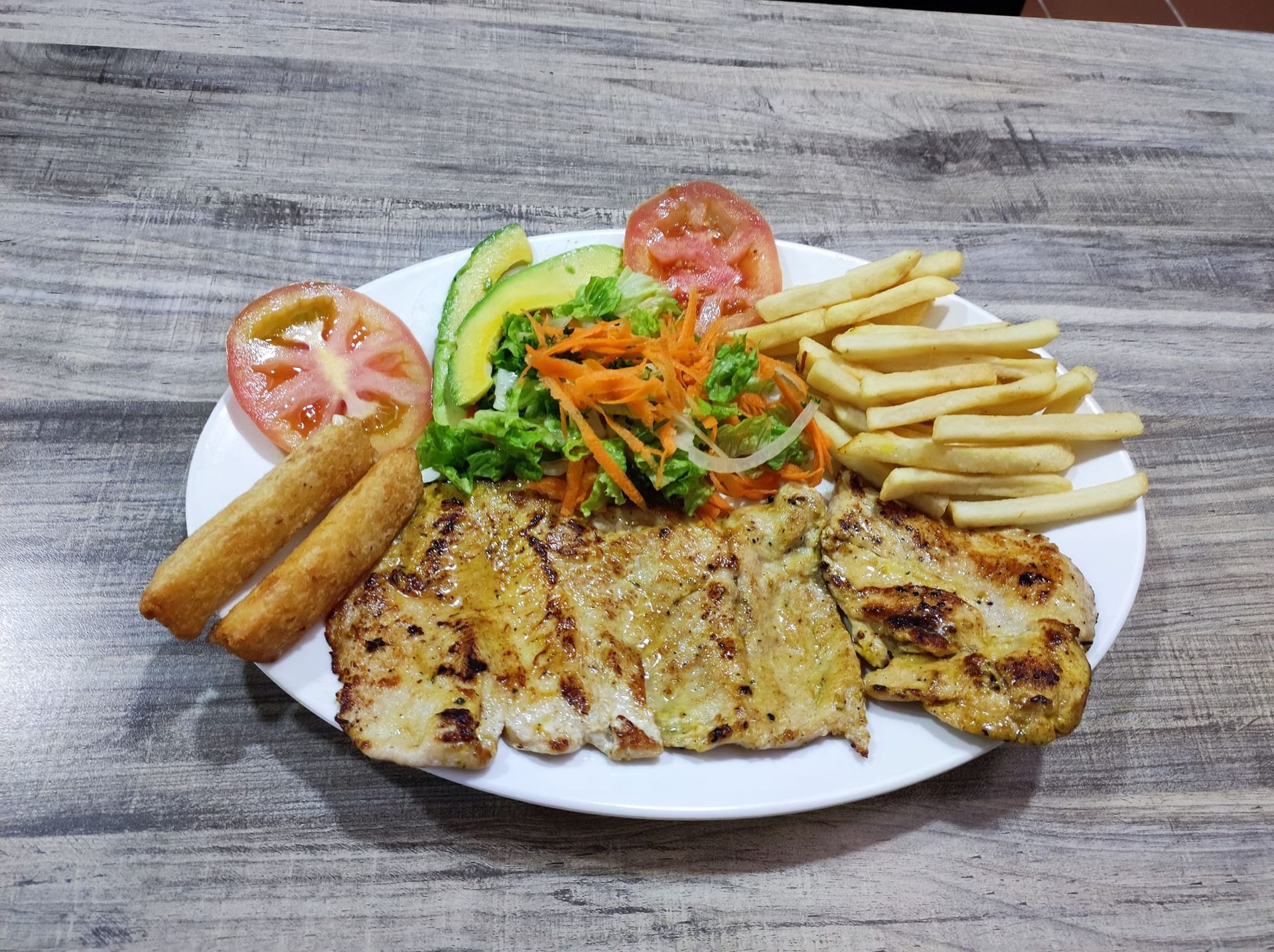 Pechuga de pollo a la parrilla con patatas fritas, ensalada, aguacate y tomate en un plato blanco.