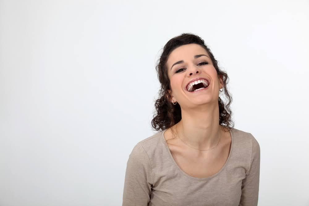 Woman with dark curly hair laughing, head tilted back, against a white background.