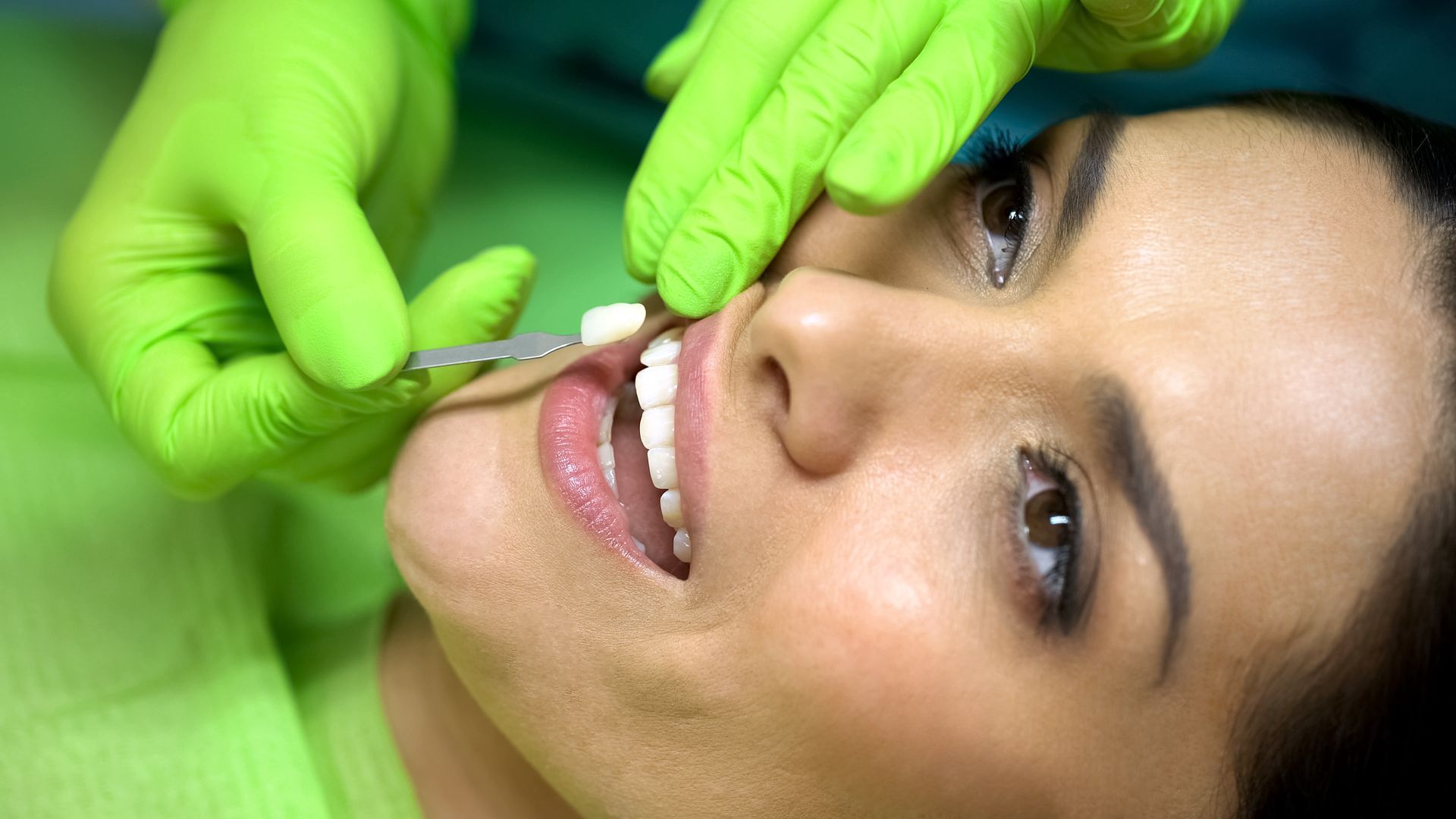 A dental professional in green gloves uses a tool to examine a patient’s teeth during a dental appointment. A dental professional in green gloves uses a tool to examine a patient’s teeth during a dental appointment.