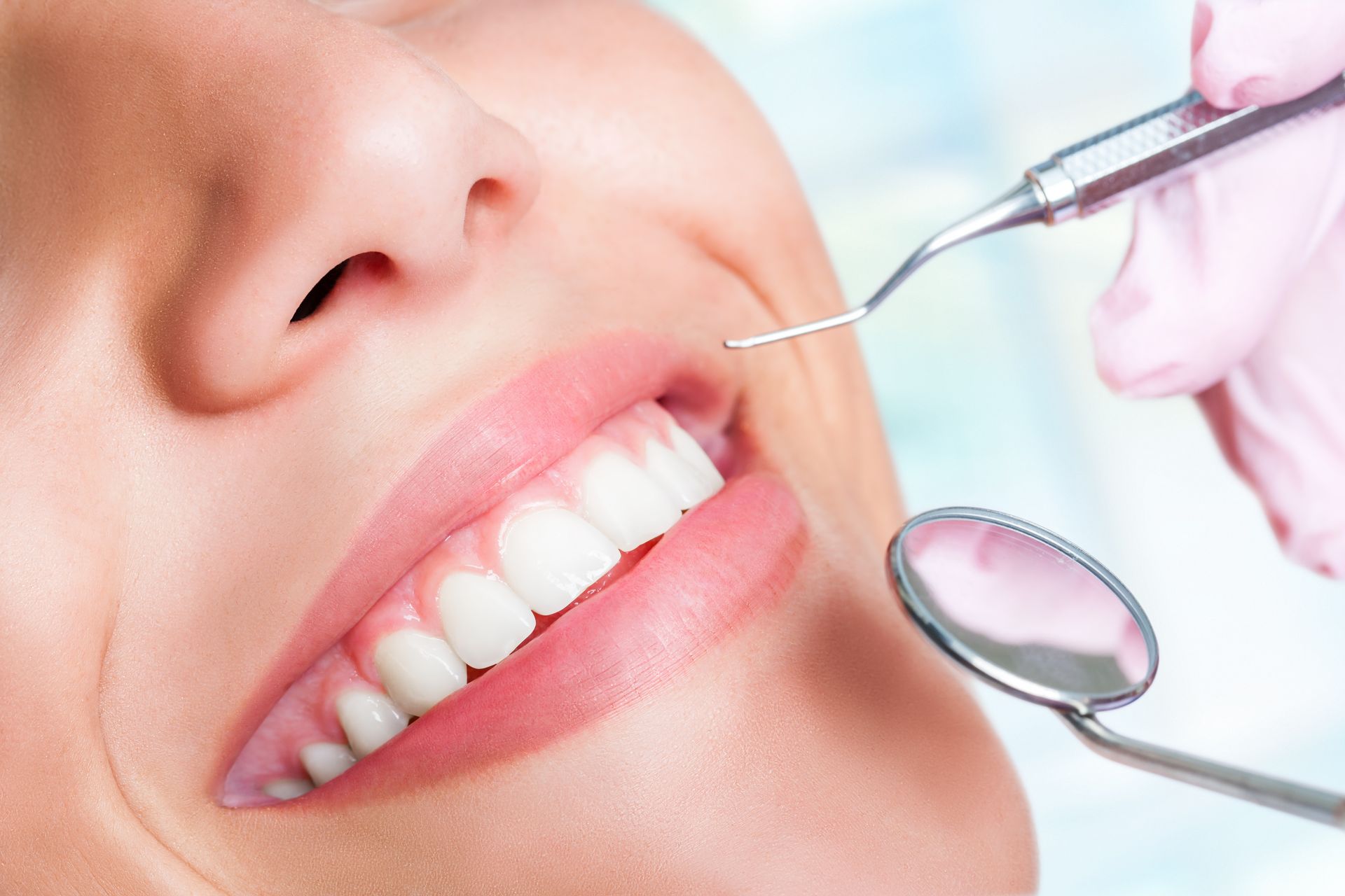 Dentist's tools examining a smiling person's healthy teeth. Pink gloves, mirror, and probe visible.