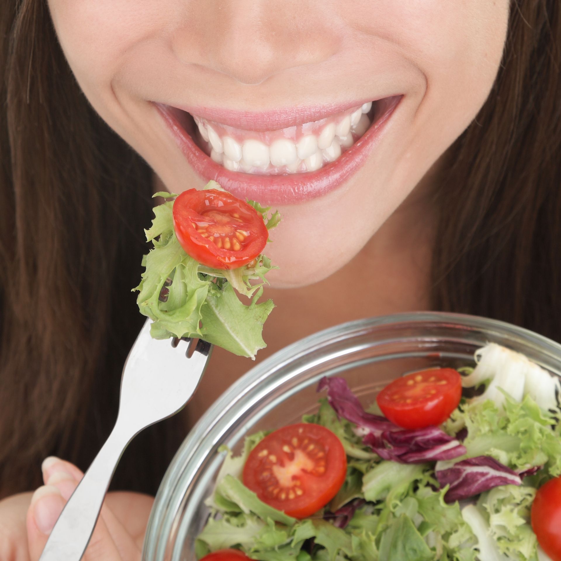 Woman smiling while eating a salad with tomatoes using a fork from a glass bowl.