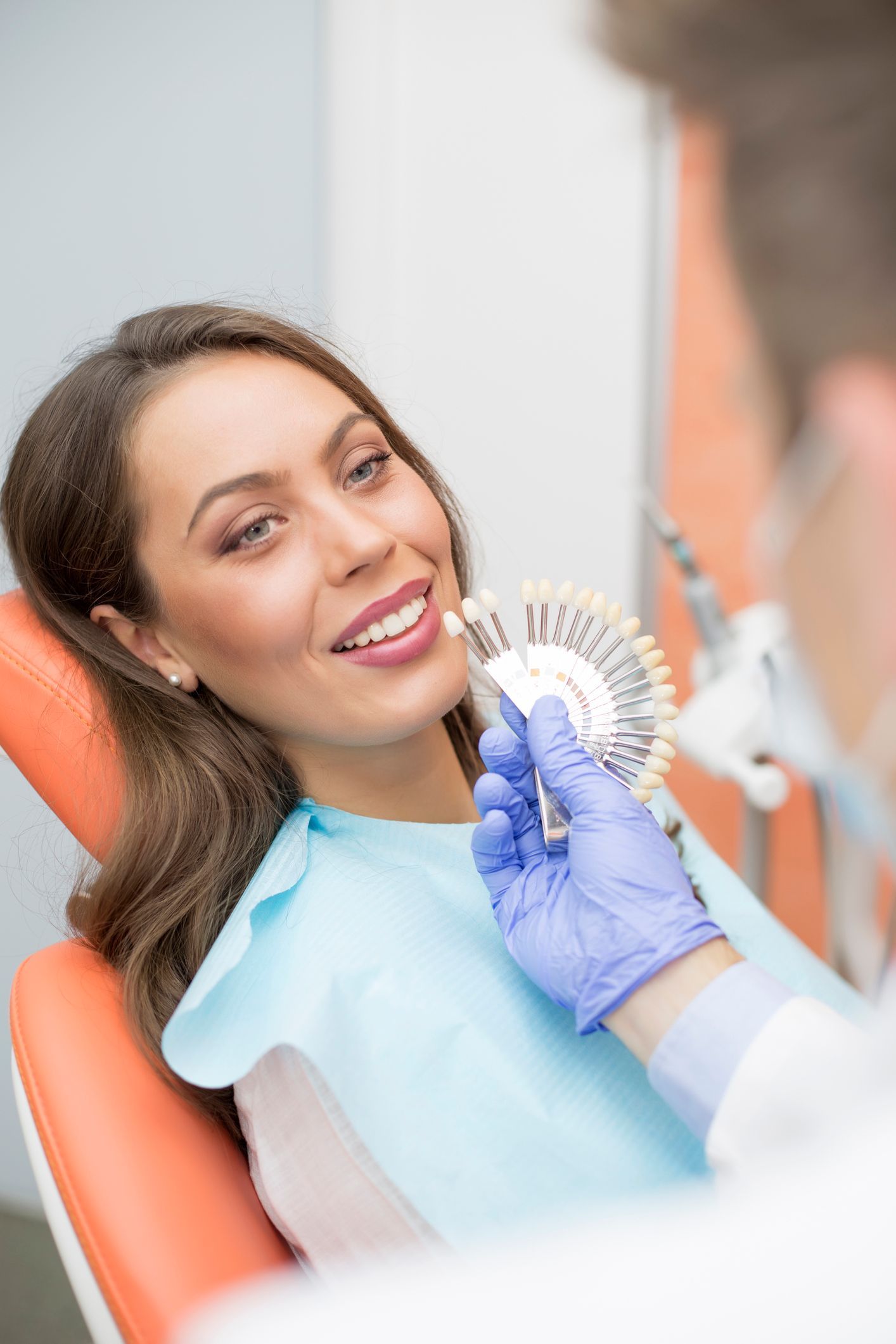 Woman in dental chair, shade matching for teeth whitening. Dentist holds a color chart.