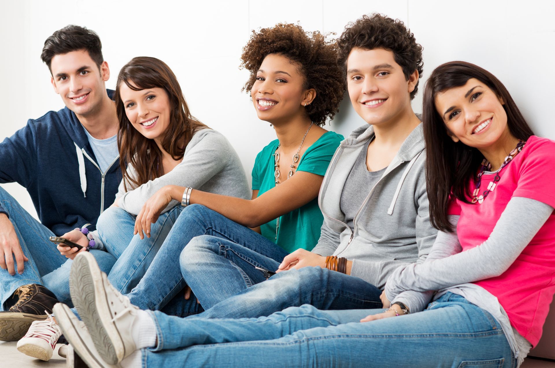 Five smiling people seated against a wall, wearing jeans and casual tops.