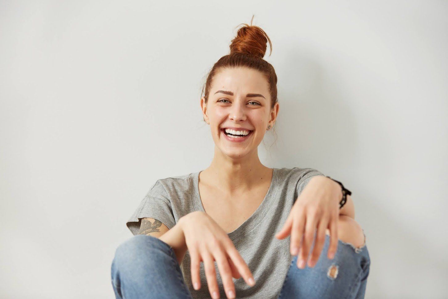 Woman with red hair in a bun laughs, sitting against a white wall in a gray tee and jeans.