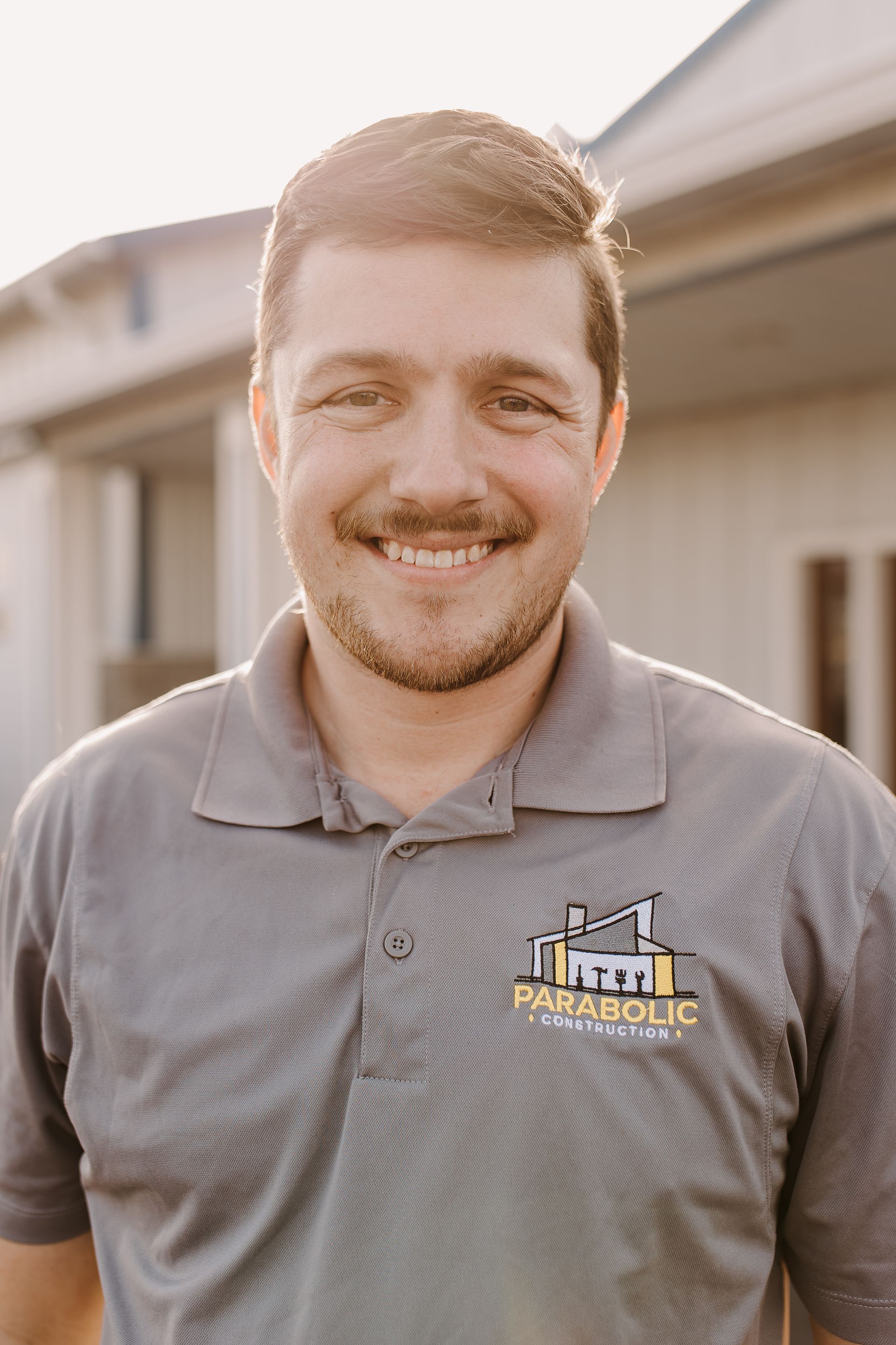 A man with a beard is wearing a gray polo shirt and smiling.