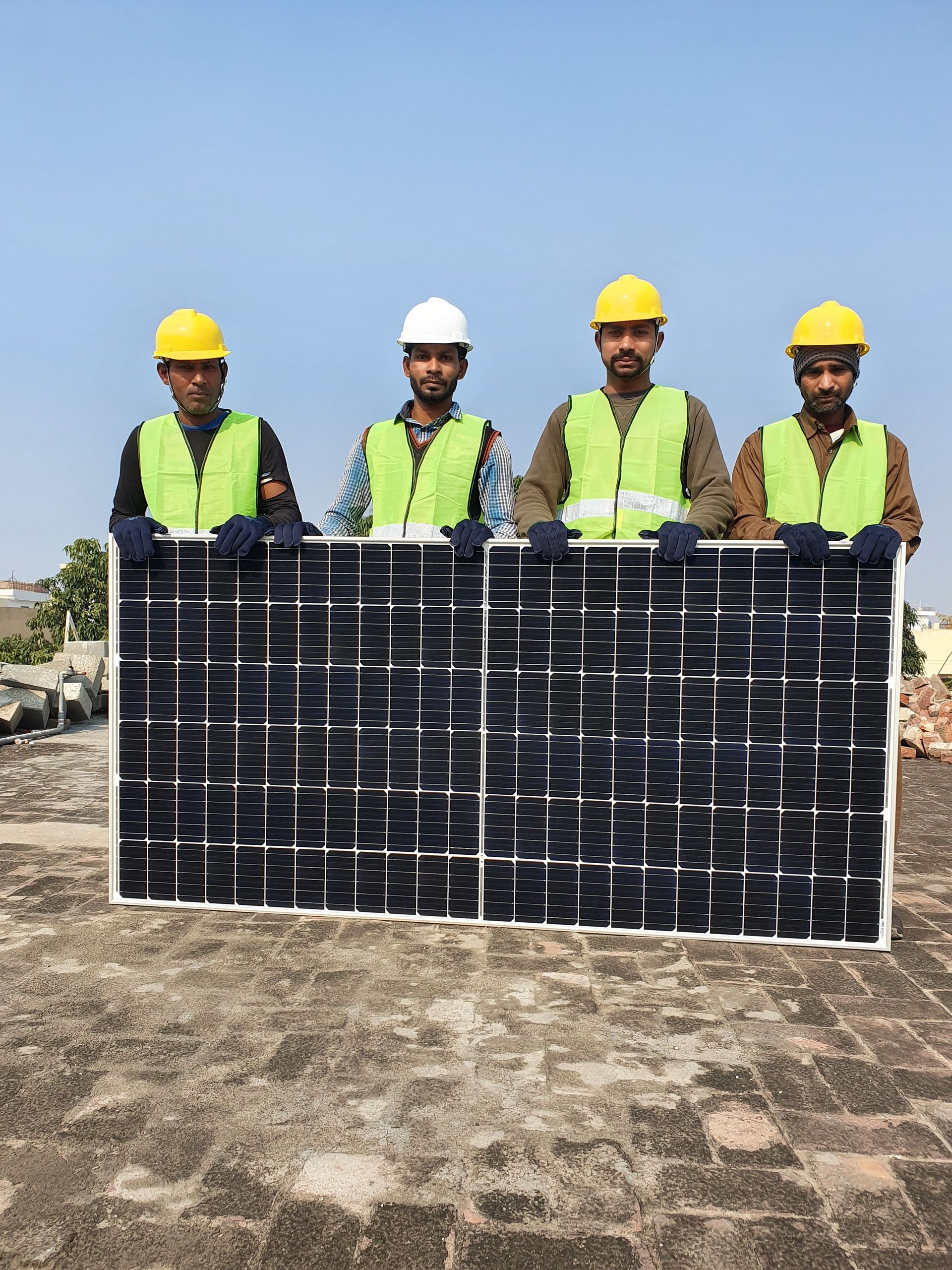Four workers in hard hats holding solar panels at a construction site under a clear sky
