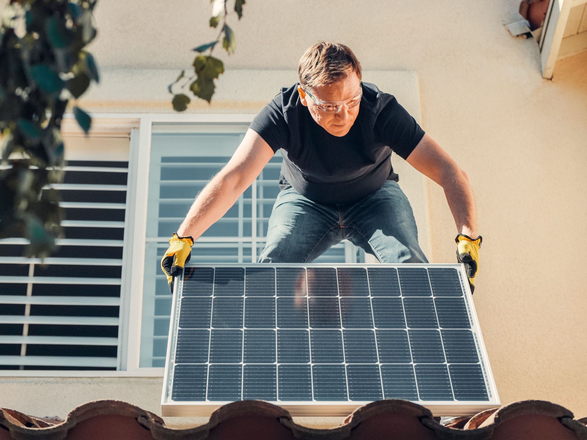 Worker installing solar panel on a rooftop, wearing gloves and leaning over the panel.