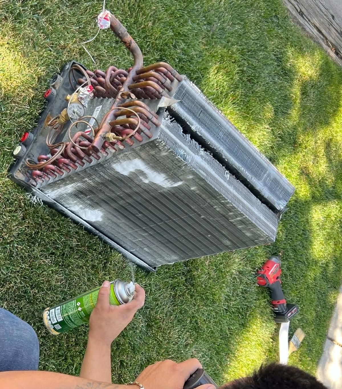 Old window air conditioner on grass, with detached copper coils and a person holding a spray can nearby