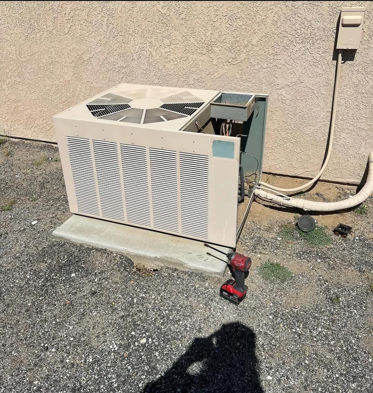 Outdoor air conditioner unit on a concrete pad beside a stucco wall, with a red drill nearby.