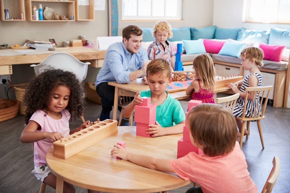 A group of children are sitting at a table in a classroom with a teacher.
