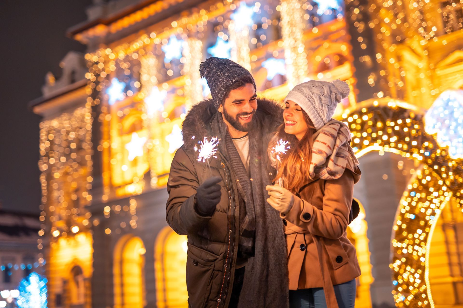 CHRISTMAS LIGHTS, Couple holding sparklers, smiling at each other in front of Christmas lights, at night.