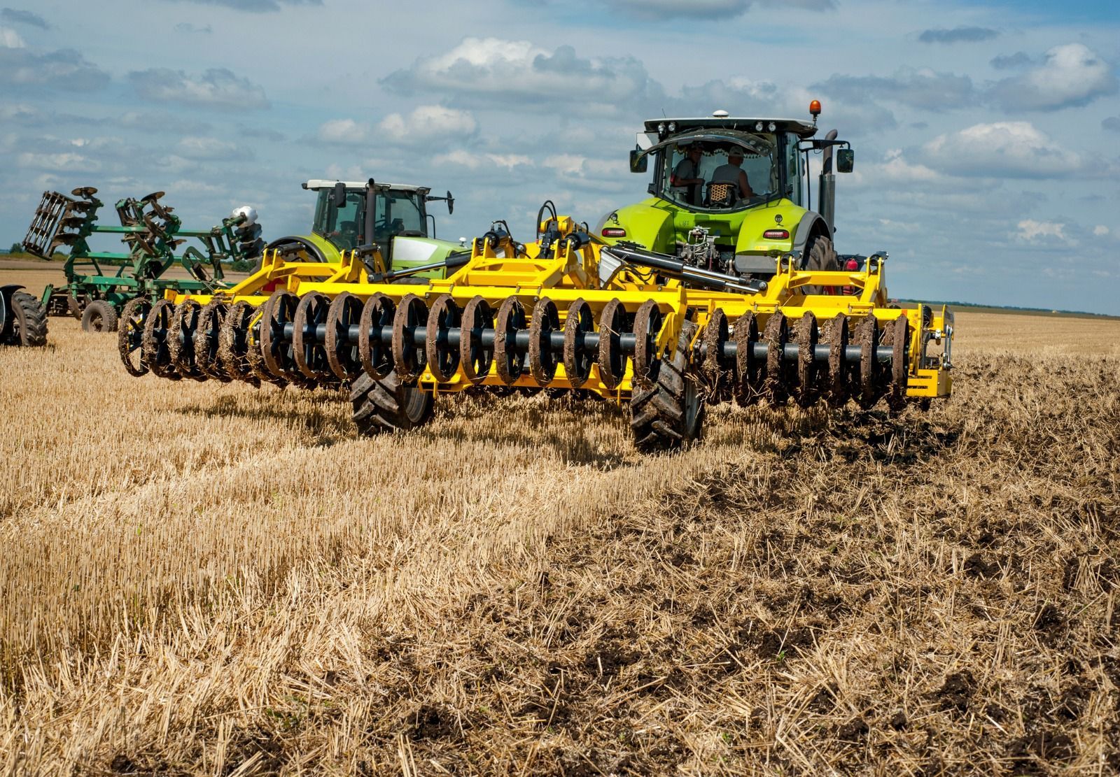 A yellow tractor is plowing a field next to a green tractor.