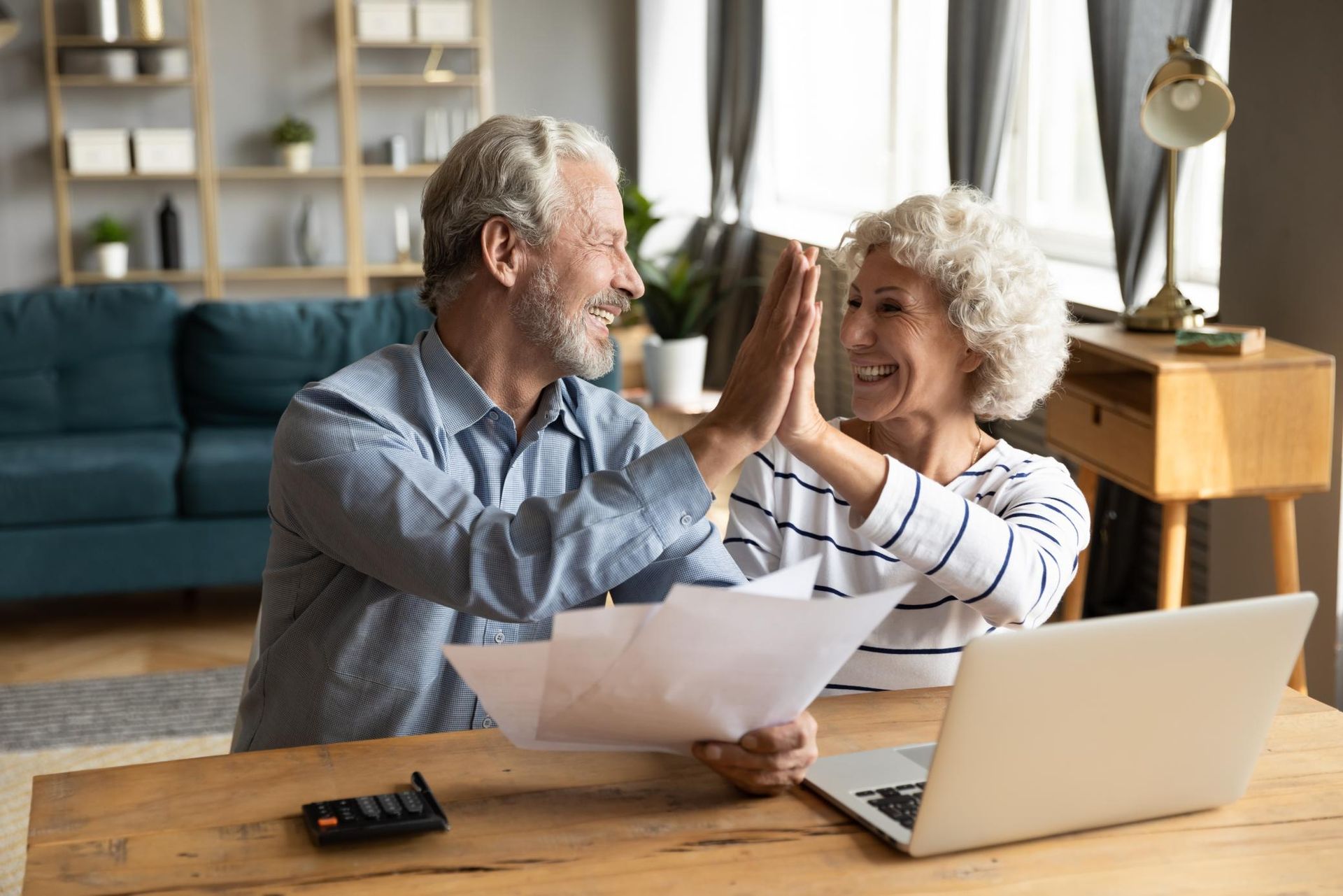 Older couple high-fiving over financial papers and a laptop in a living room.