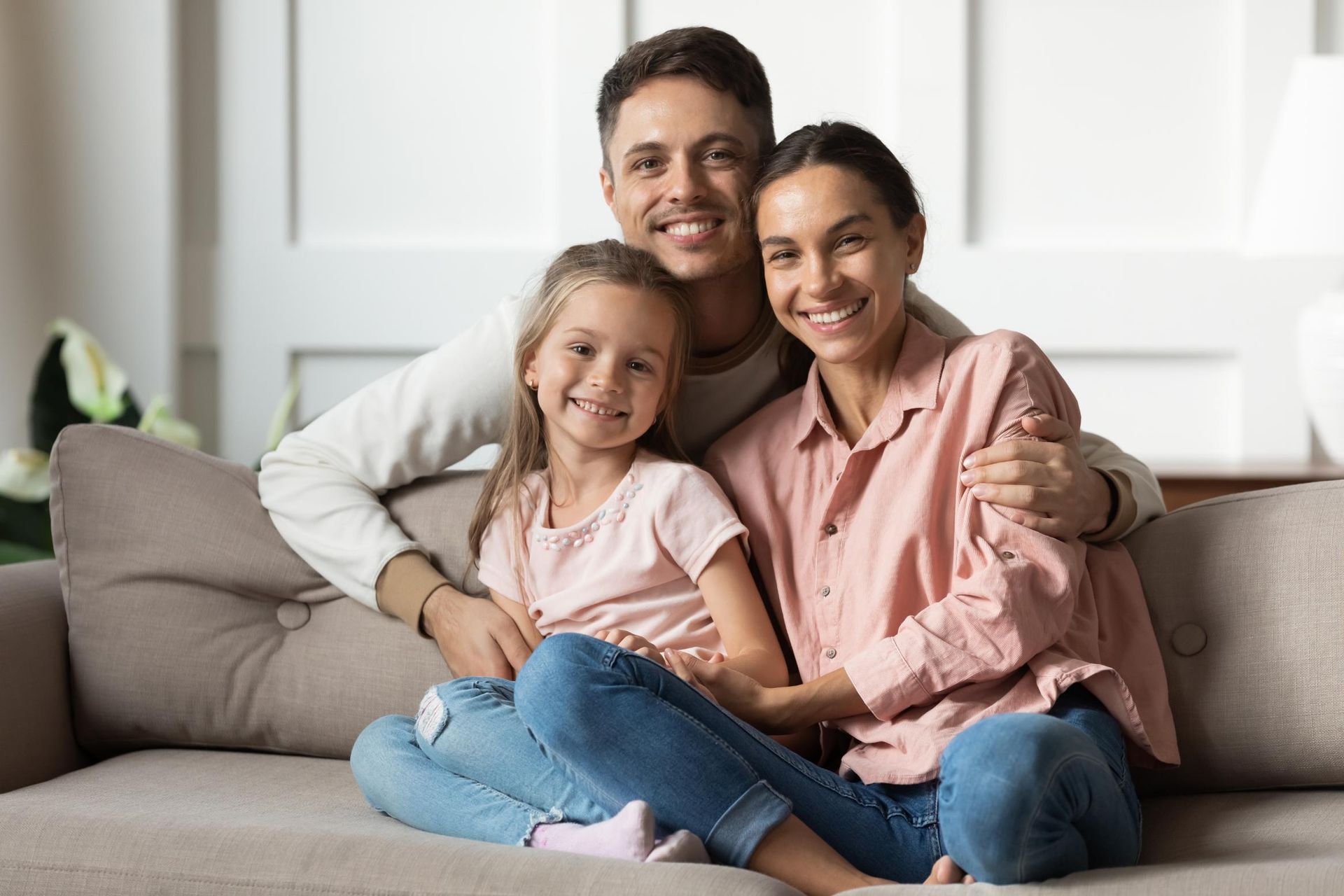 Family smiling on a couch: man has arm around woman, child sits in front. Warm tones.