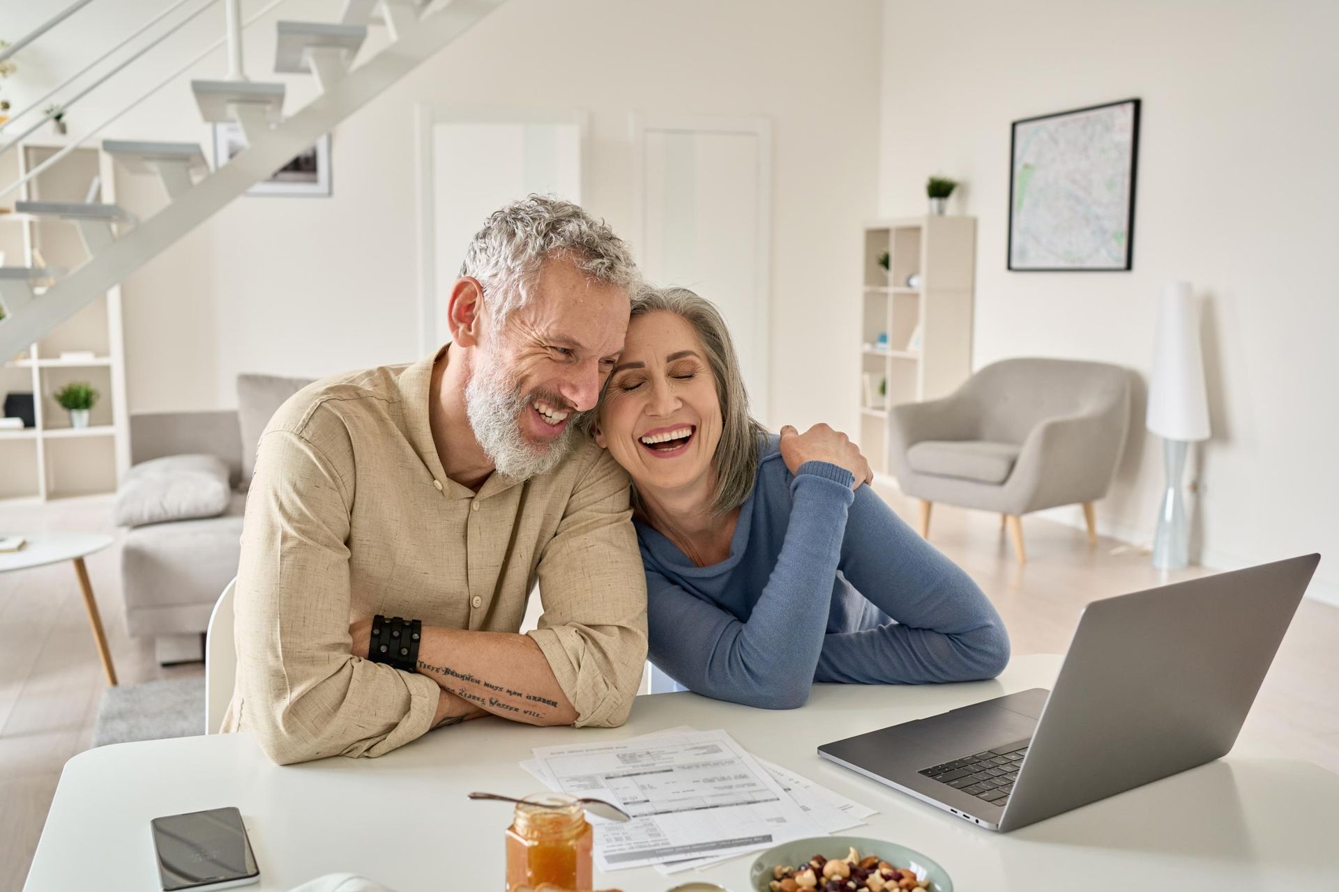 Couple laughing, looking at laptop and paperwork at a table in a bright, modern living room.