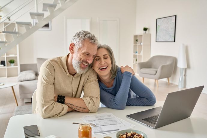 Couple laughing, looking at laptop and paperwork at a table in a bright, modern living room.