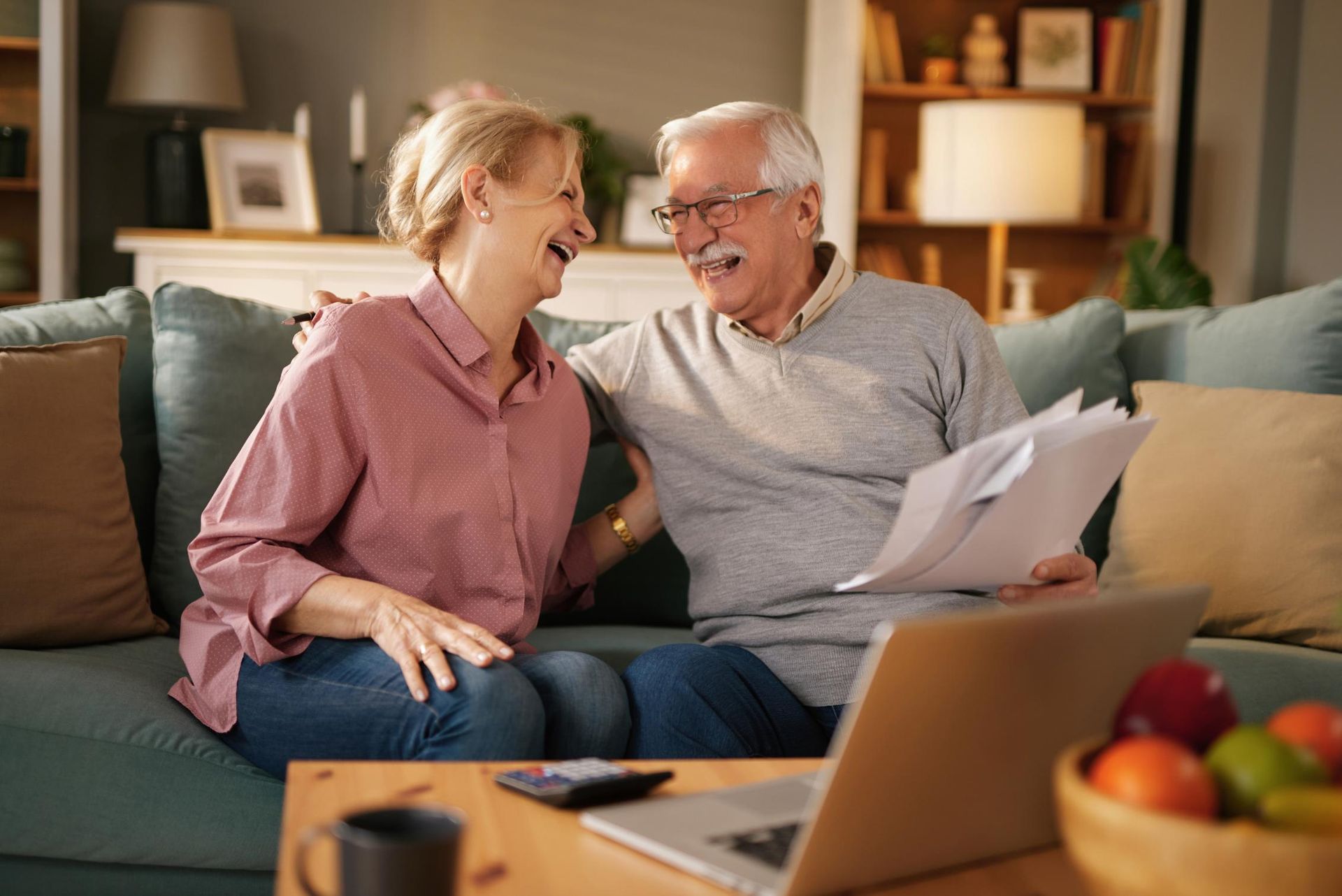 Smiling couple on a sofa, looking at papers and laptop in a living room.