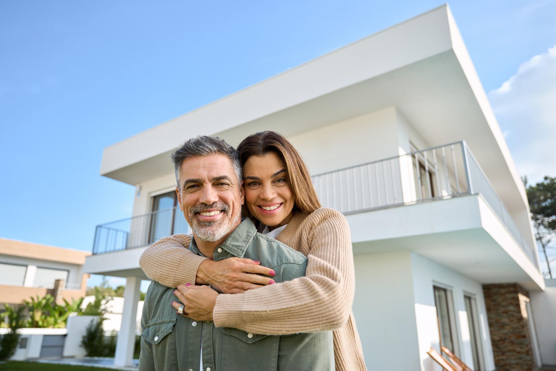 Couple smiling in front of a modern white house, woman hugging man. Blue sky.