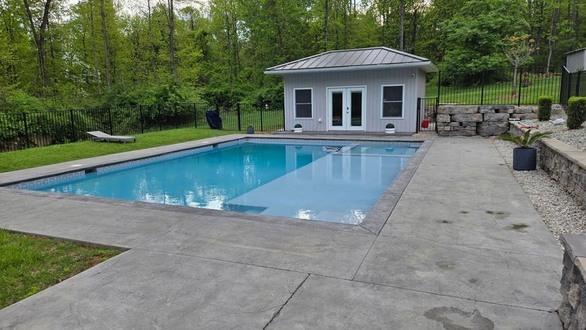 Backyard swimming pool with a concrete patio and a small gray pool house surrounded by trees.