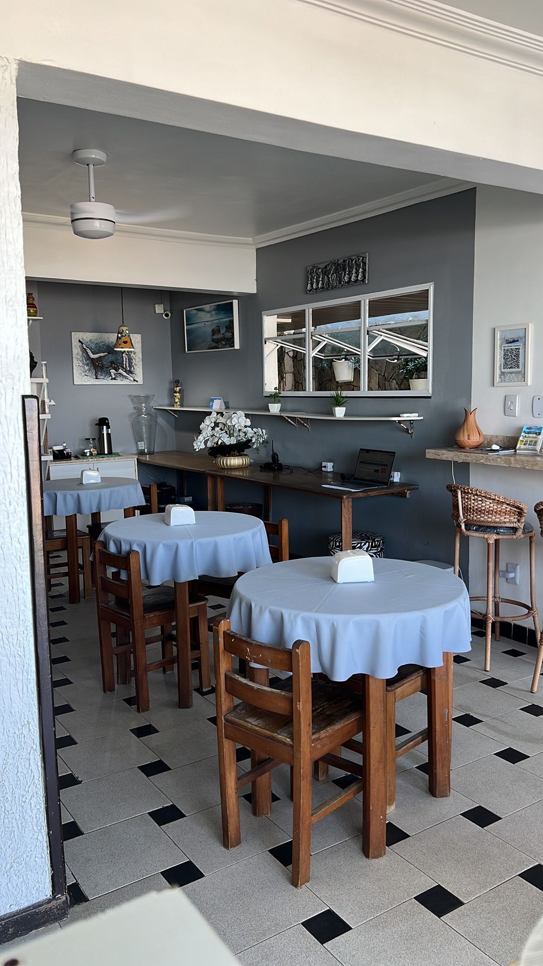 Interior do café: mesas redondas com toalhas de mesa azuis, cadeiras de madeira, piso xadrez.