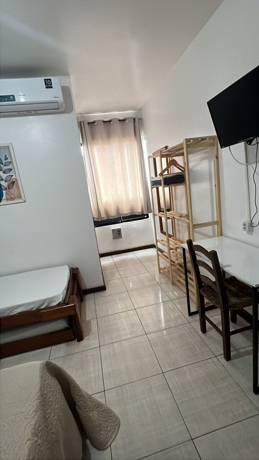 Quarto pequeno com cama, estante e escrivaninha. Paredes brancas, piso de azulejo e TV.