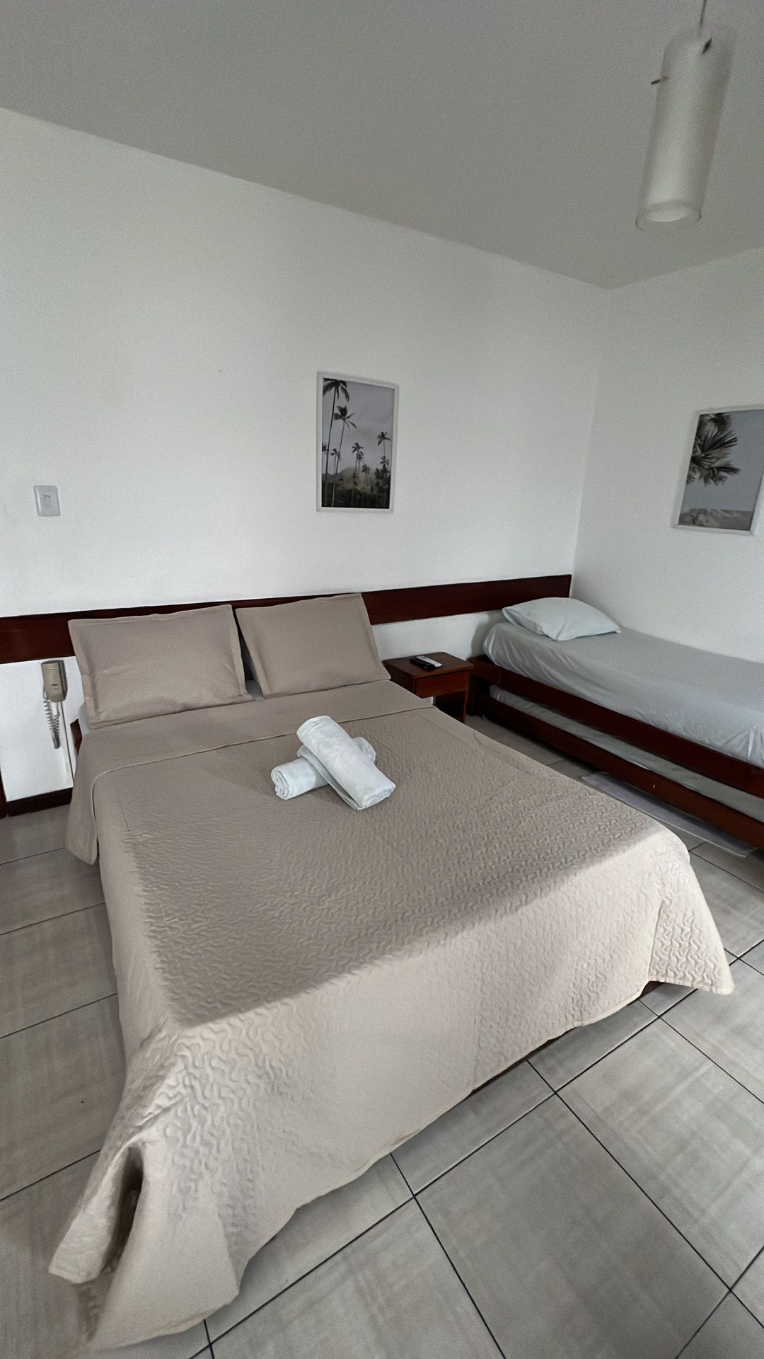 Quarto com duas camas, roupa de cama bege, paredes brancas, dois pequenos quadros e piso de azulejo.