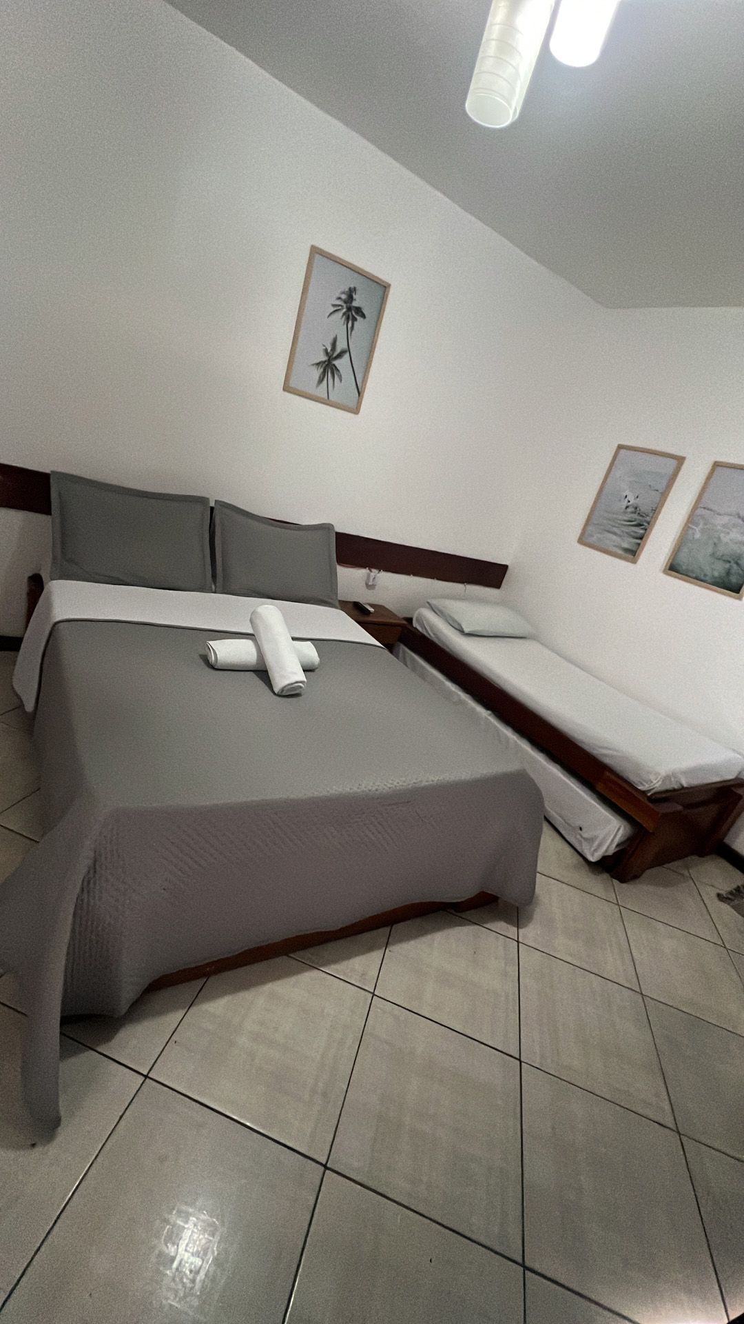 Quarto com uma cama de casal e uma cama de solteiro. Roupa de cama cinza, piso de azulejo, obras de arte na parede.