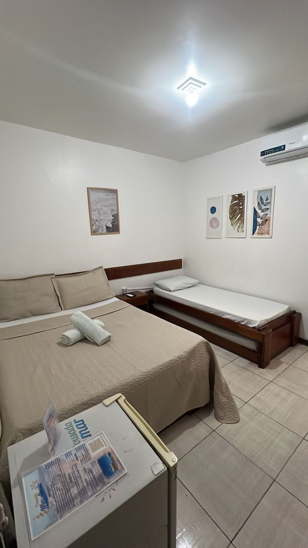 Quarto com cama de casal, cama de solteiro e frigobar. Paredes brancas, decoração com obras de arte.