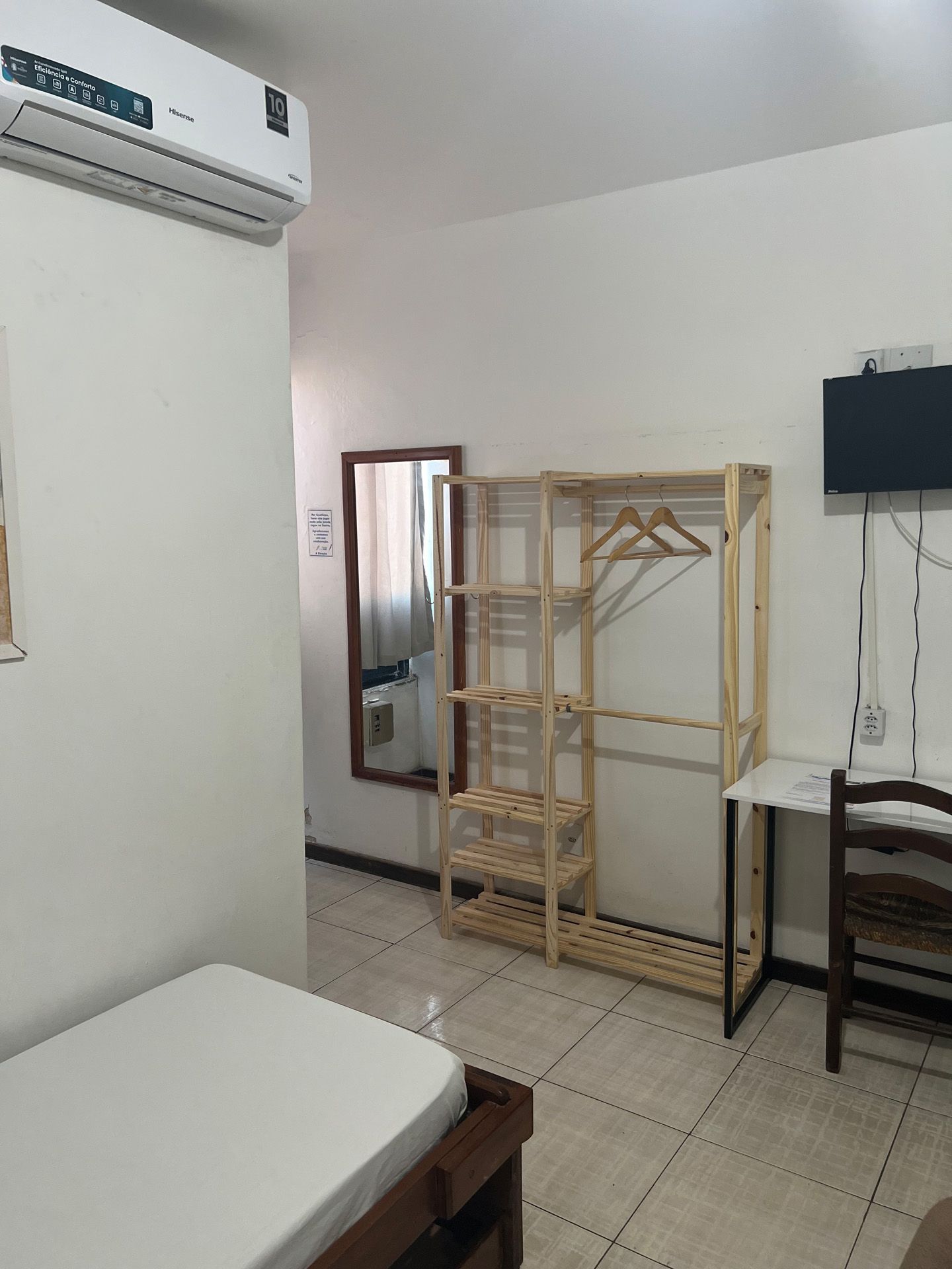 Quarto pequeno com cama, estante de madeira, espelho, escrivaninha com cadeira e ar condicionado.