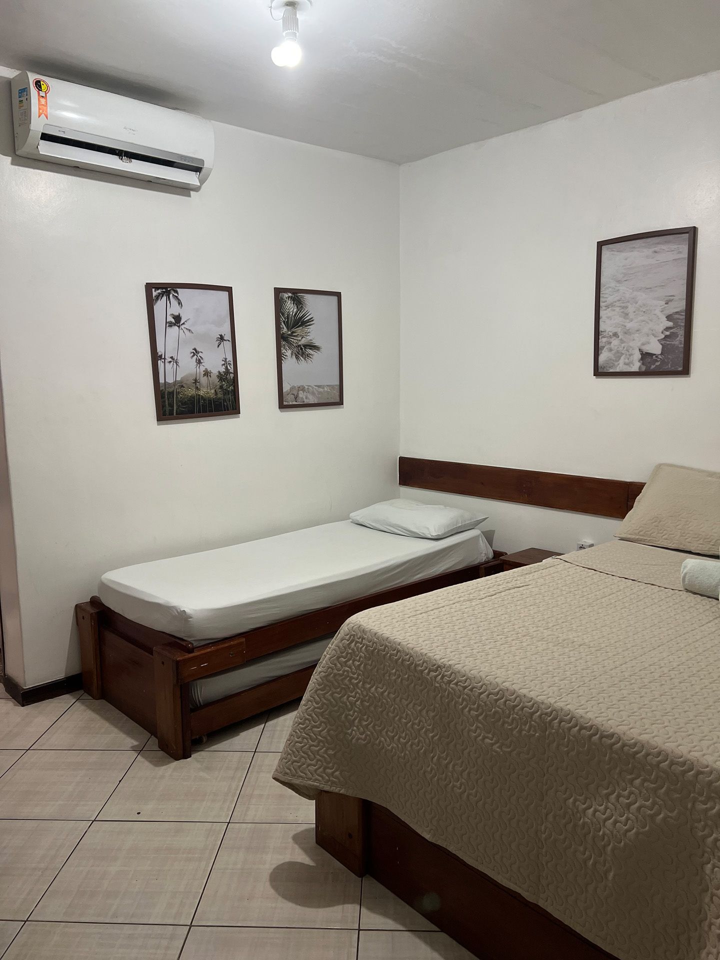 Quarto com duas camas de madeira, duas obras de arte emolduradas, ar condicionado e luminária.