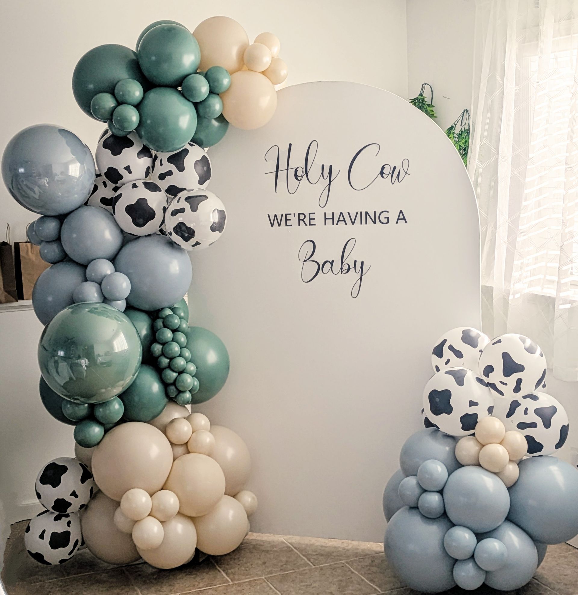 A baby shower backdrop featuring a balloon arch in blue, cream, and cow print with the text 