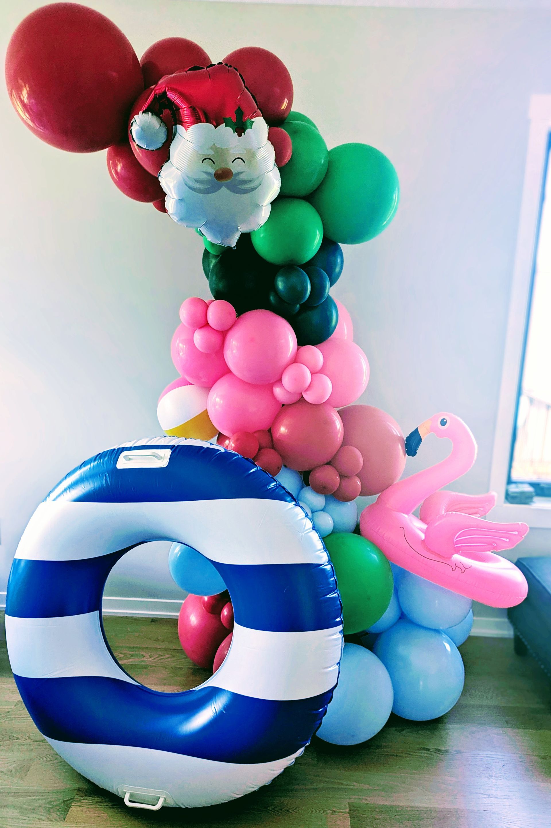 A whimsical balloon display featuring a Santa face, flamingo, and beach ring in a colorful arch against a wall.