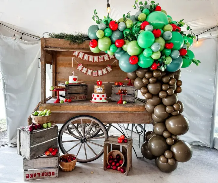 A dessert table decorated like an apple cart. Green, red, and brown balloons form an arch and tree-like display.