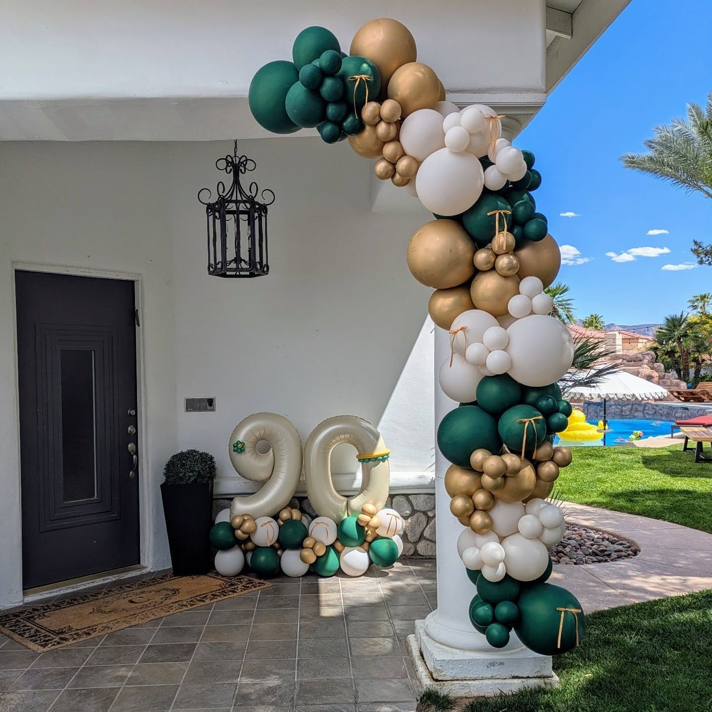 A decorated entrance for a 90th birthday. Green, gold, and white balloon arch, with large foil “90” balloons.