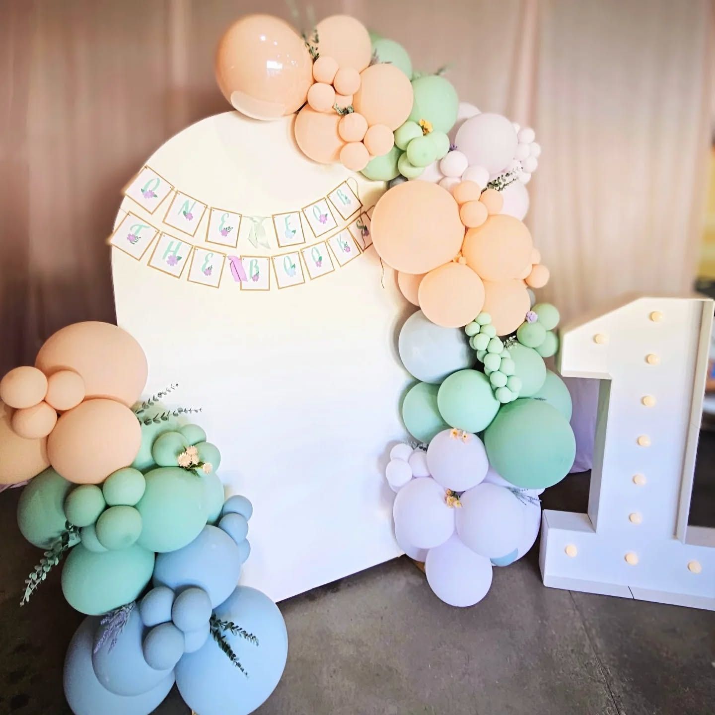 A celebratory first birthday display with a white arch, pastel-colored balloon garland, banner, and illuminated number 