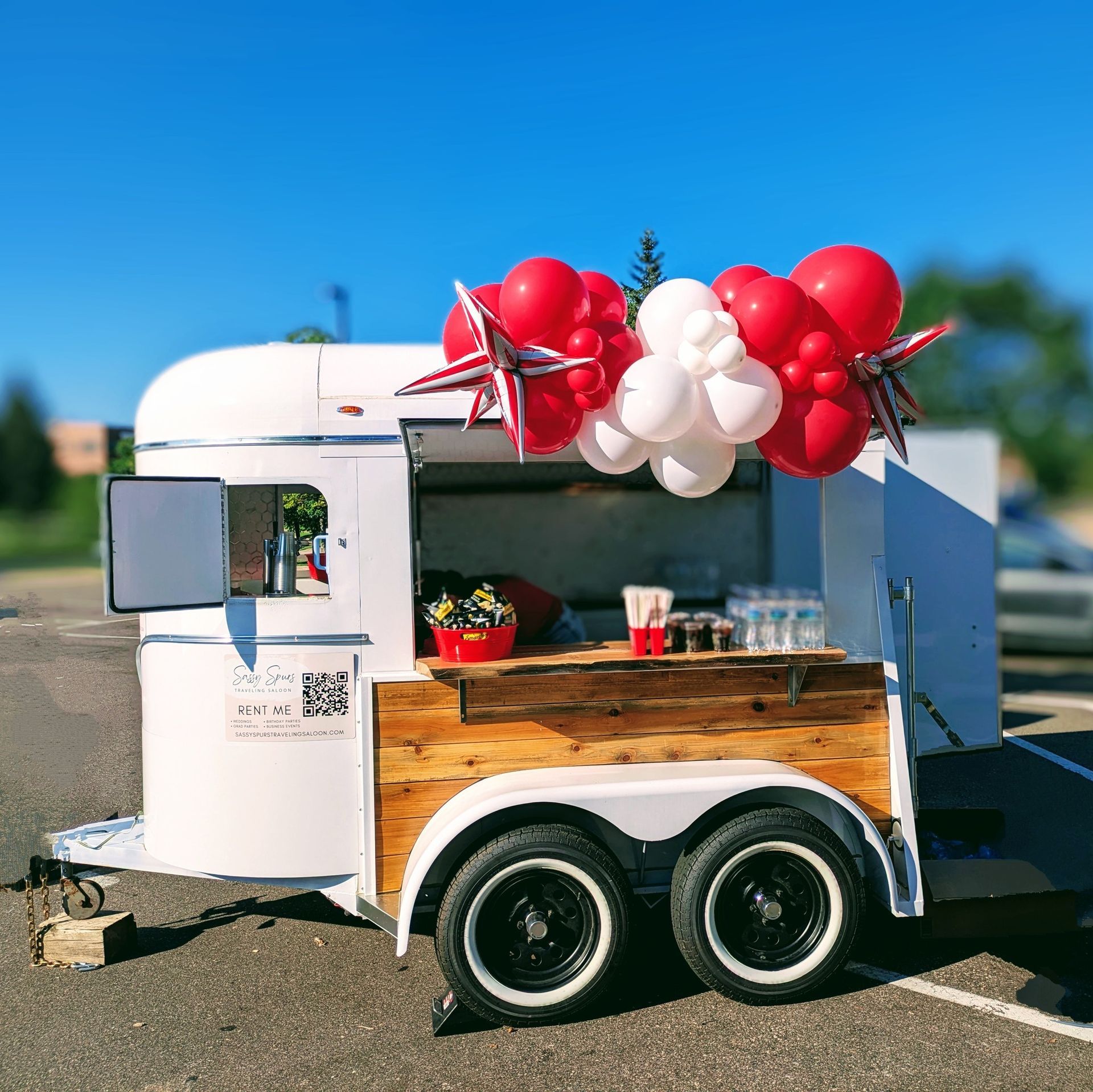 A repurposed white horse trailer with a wooden counter serving drinks. Red, white, and star-shaped balloons decorate the top.