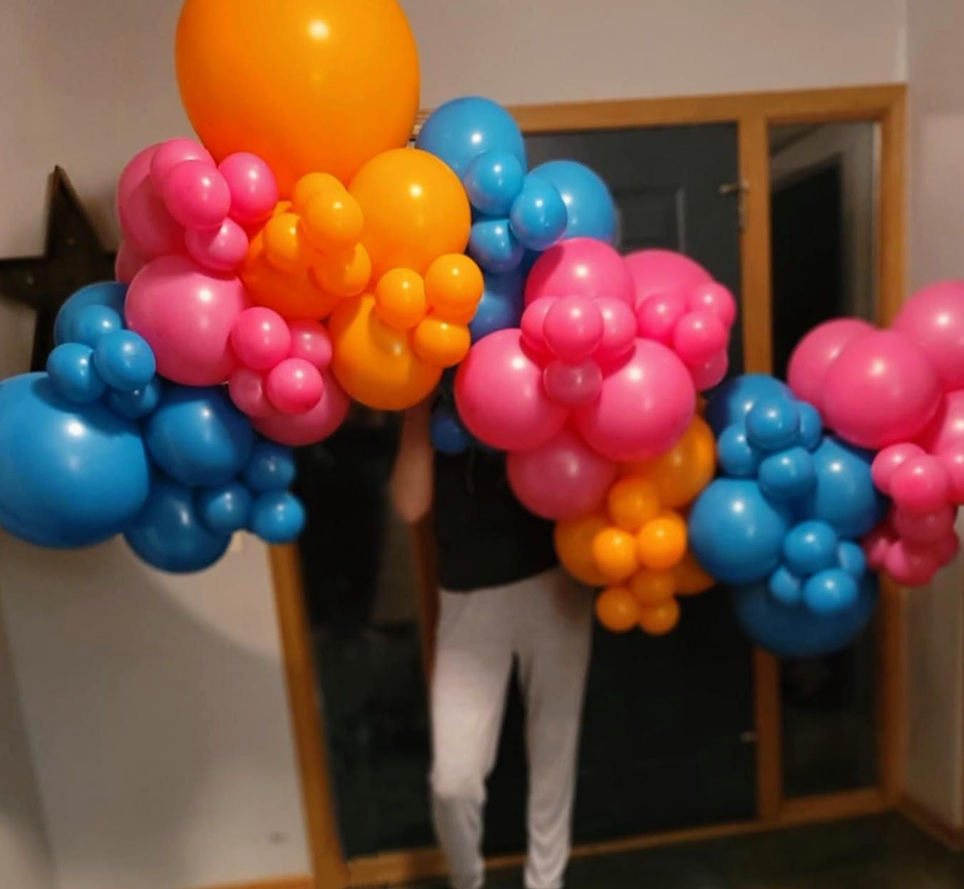 Person holding a colorful balloon arch with pink, orange, and blue balloons in front of a doorway.
