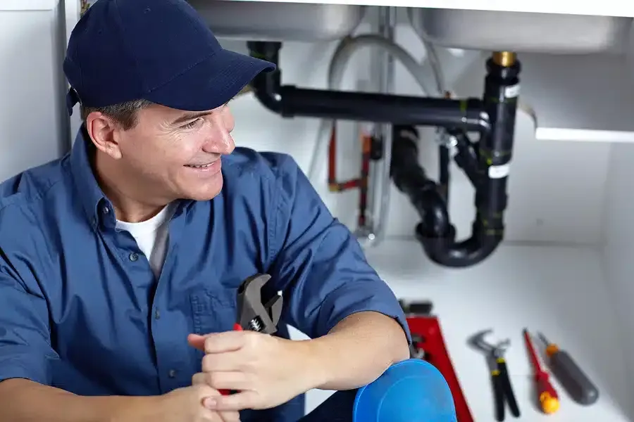 Plumber smiling, holding wrench, under a sink, tools visible.
