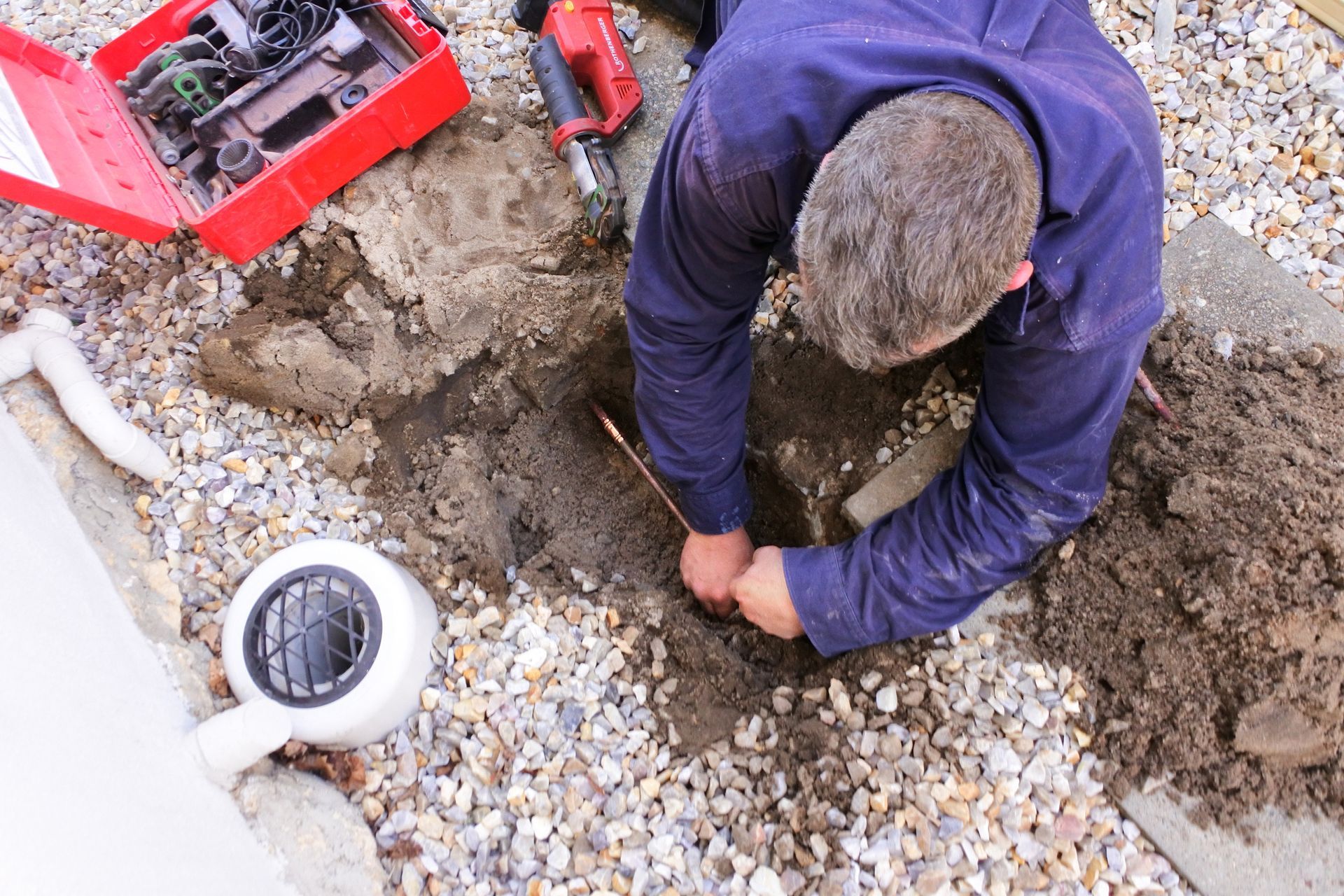 A person in a blue uniform digs in gravel, working on an underground pipe near a red tool case.