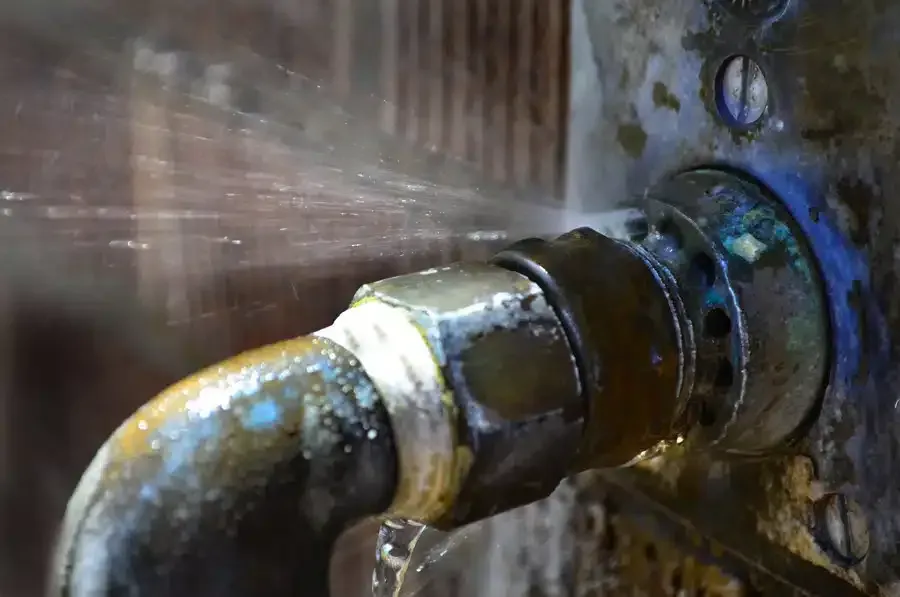 Leaking pipe joint with water spraying outwards; metal, corrosion, close-up.