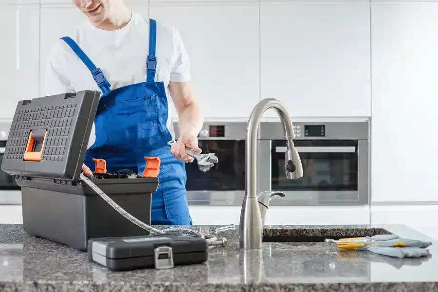 Plumber in blue overalls working on a kitchen sink with tools.