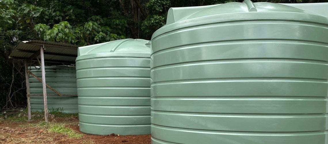 Three green water tanks lined up, with a small covered structure on the left, surrounded by greenery.