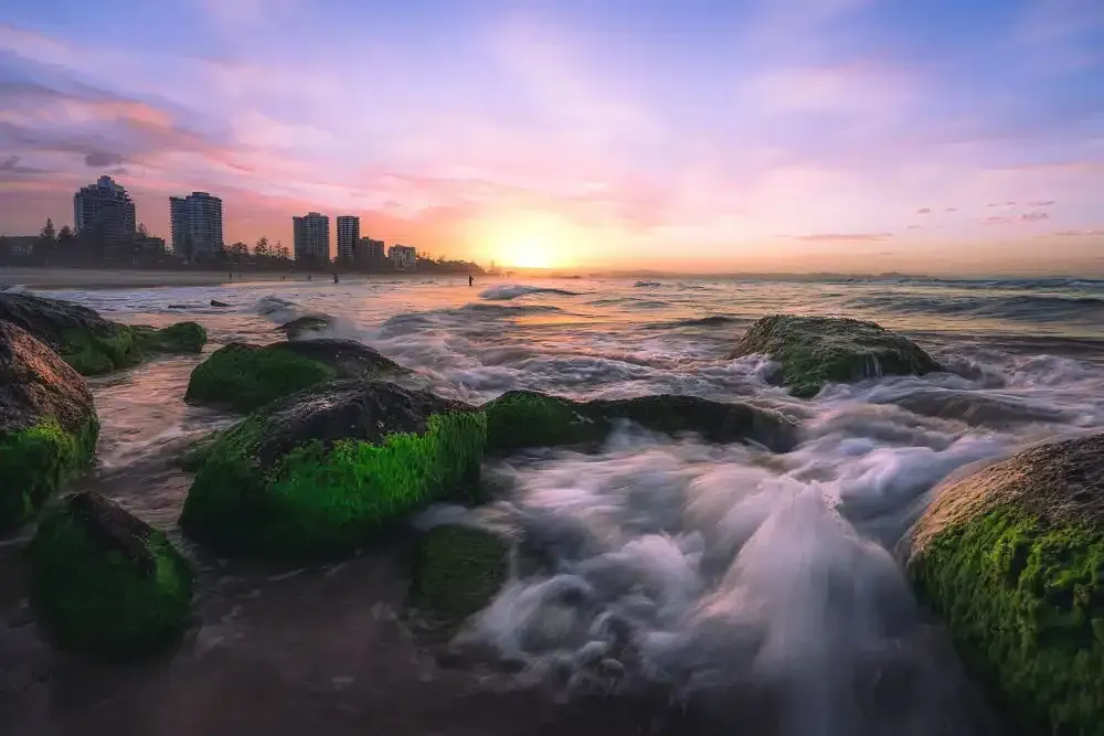 Sunset over ocean, waves crashing on green-covered rocks, silhouetted city skyline in distance.